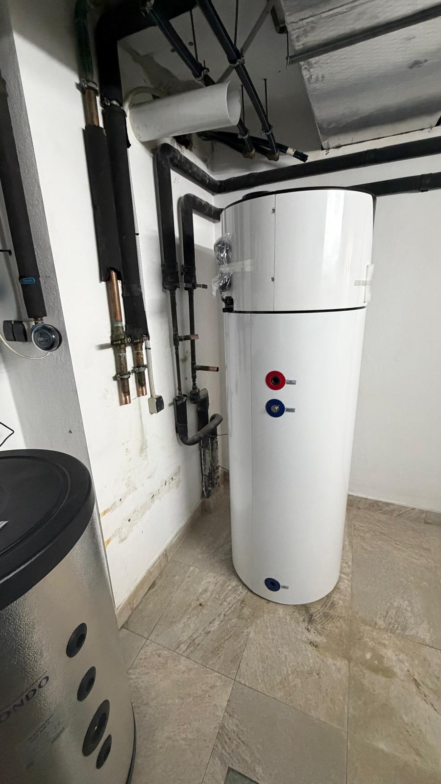 Side view of Daitsu aerothermal heat pump installed in La Cala de Mijas plant room — Redondo stainless steel buffer cylinder to left, new black insulated pipework and distribution manifold visible
