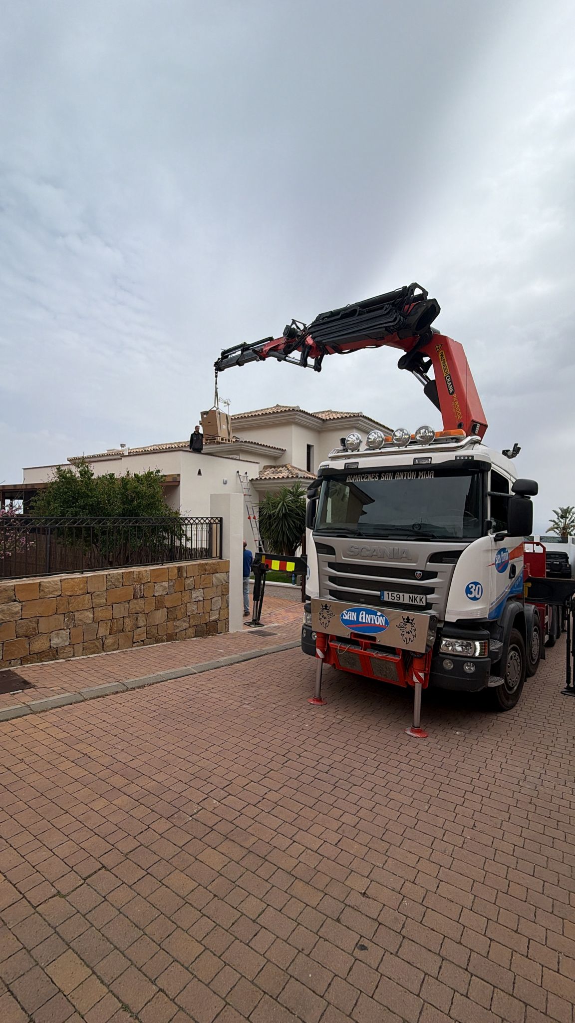 Front view of Scania San Antón crane truck during heat pump delivery at La Cala de Mijas villa — technician on rooftop receiving outdoor unit during professional Agua Therm installation