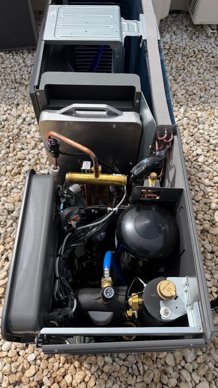 Interior of Midea heat pump outdoor unit — black spherical compressor, copper refrigerant circuit pipework, brass valves and electrical wiring visible with panel open during installation at La Cala de Mijas