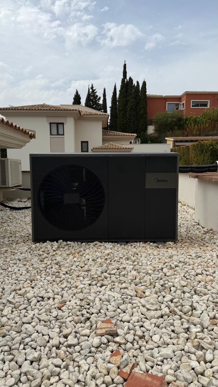Midea heat pump outdoor unit installed on flat roof of La Cala de Mijas villa — black unit, front fan grille, Midea logo visible, neighbouring villa rooftops and cypress trees in background