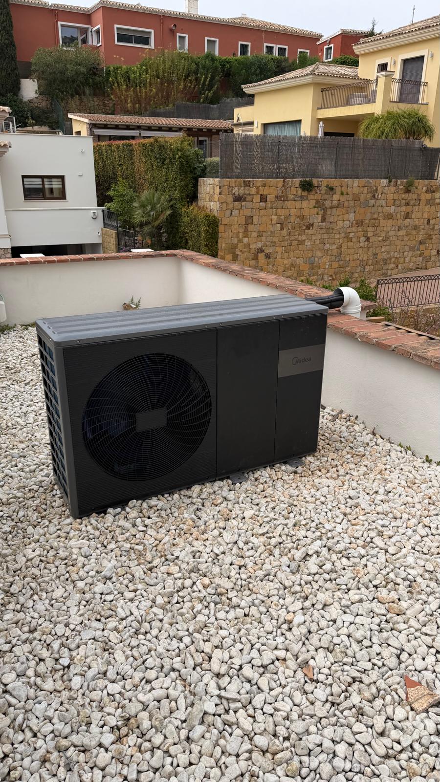 Side angle view of Midea heat pump outdoor unit on La Cala de Mijas villa rooftop — black connection pipework visible on side, neighbouring villas and trees in background, overcast sky
