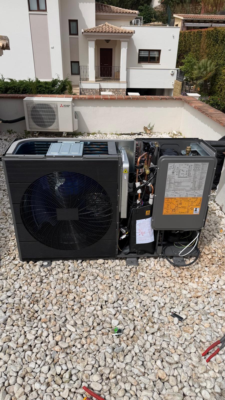 Midea heat pump outdoor unit with panel open during commissioning on La Cala de Mijas rooftop — refrigerant circuit visible, condenser coil, wiring, Mitsubishi outdoor unit on neighbouring property in background