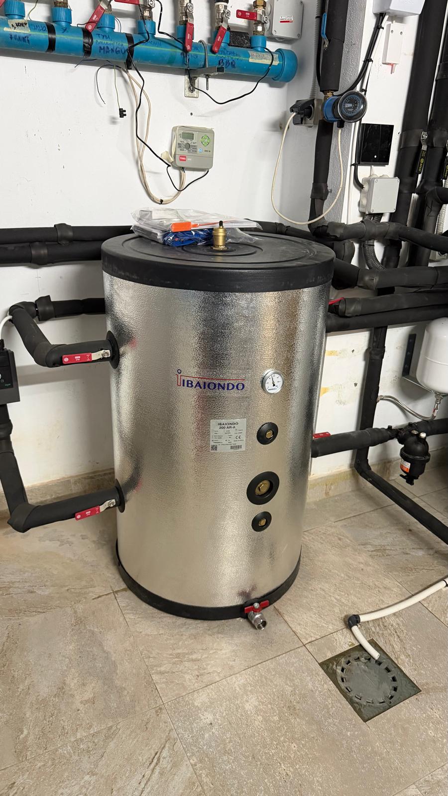 Ibaiondo stainless steel inertia buffer tank installed in La Cala de Mijas plant room — Ibaiondo label visible, inlet and outlet connections with red isolation valves, sediment filter and distribution manifold in background