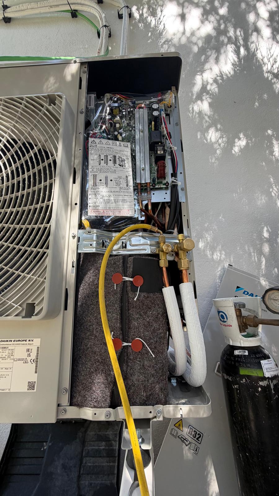 Unidad exterior Daikin con tapa retirada mostrando placa de control original del fabricante junto a botella de refrigerante R32
