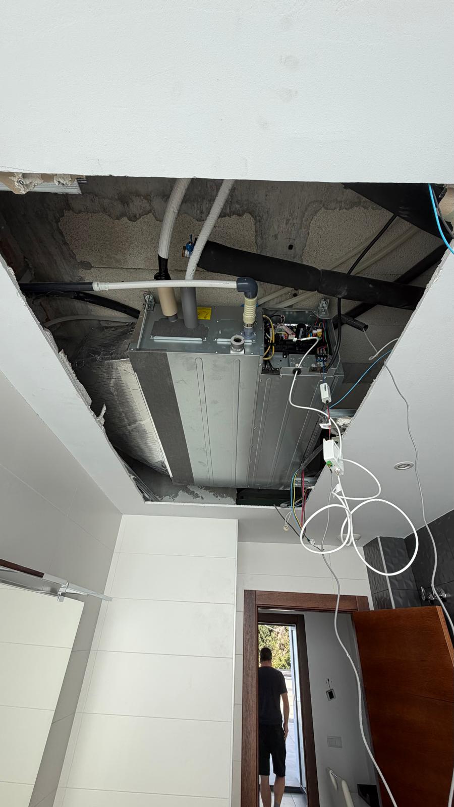 Underside view of Daikin indoor unit in ceiling void: grey unit body, condensate drain PVC pipe, Airzone control wiring, technician visible in background