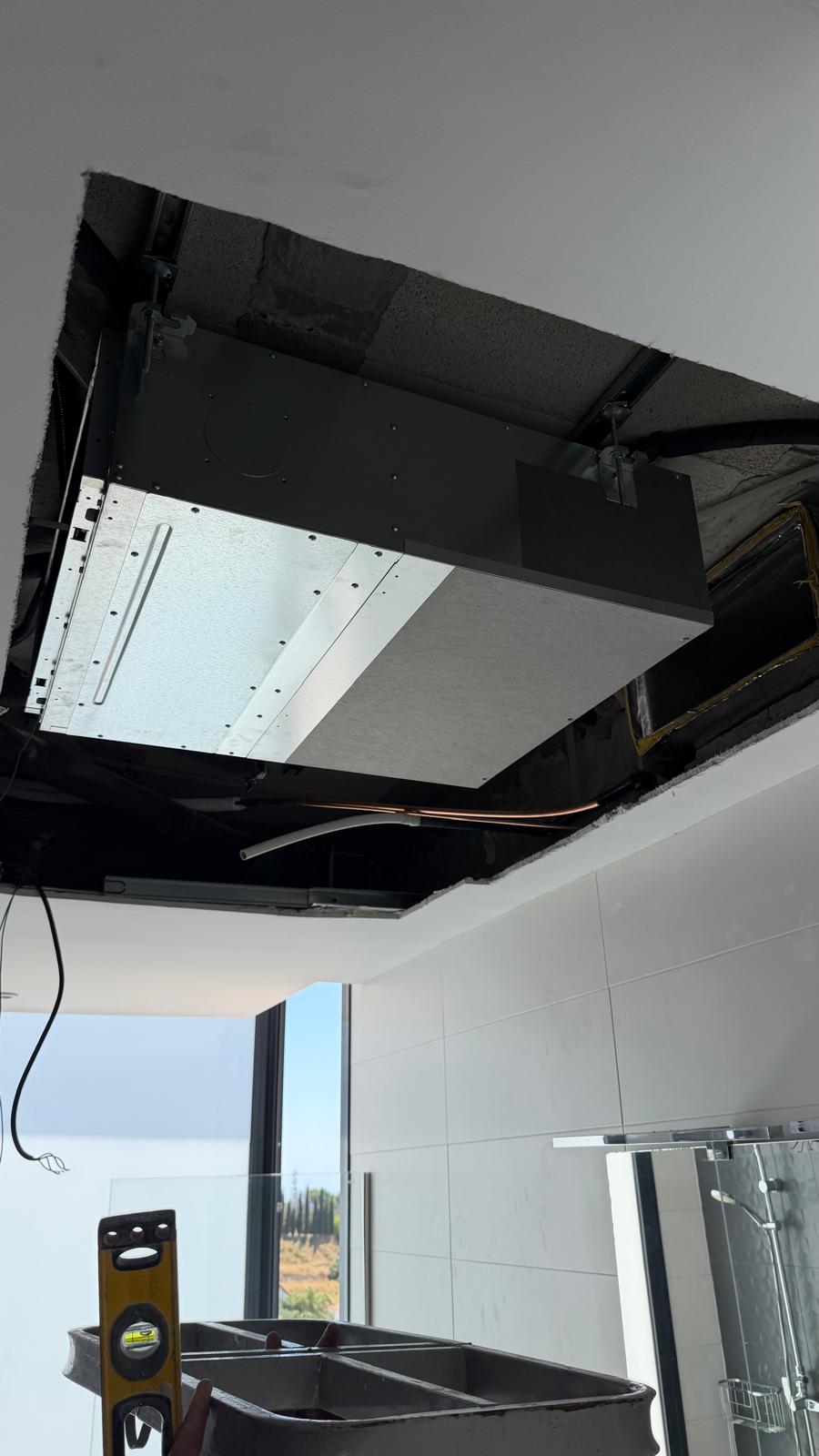 Daikin indoor unit in ceiling void close-up: copper line connection at side visible, spirit level on work surface, glass shower screen below