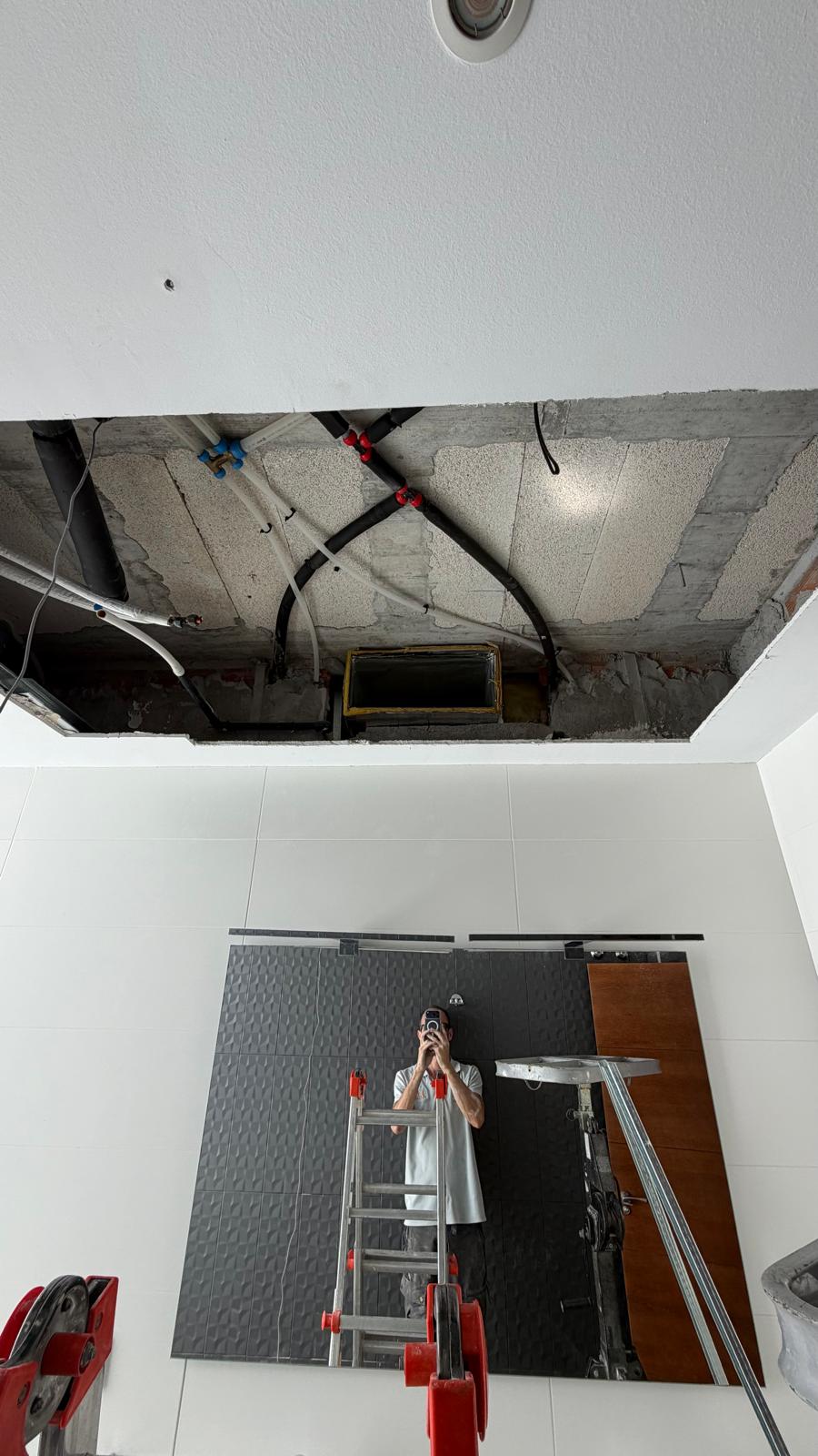 Second ceiling void: refrigerant lines routed (black insulation with blue/red valve fittings), supply grille aperture cut into ceiling, technician reflected in mirror below