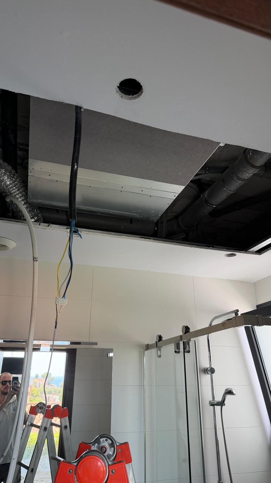 Bathroom ceiling void: unit installed with flex duct (left), electrical wiring (yellow/green, blue), corrugated conduit, glass shower screen below