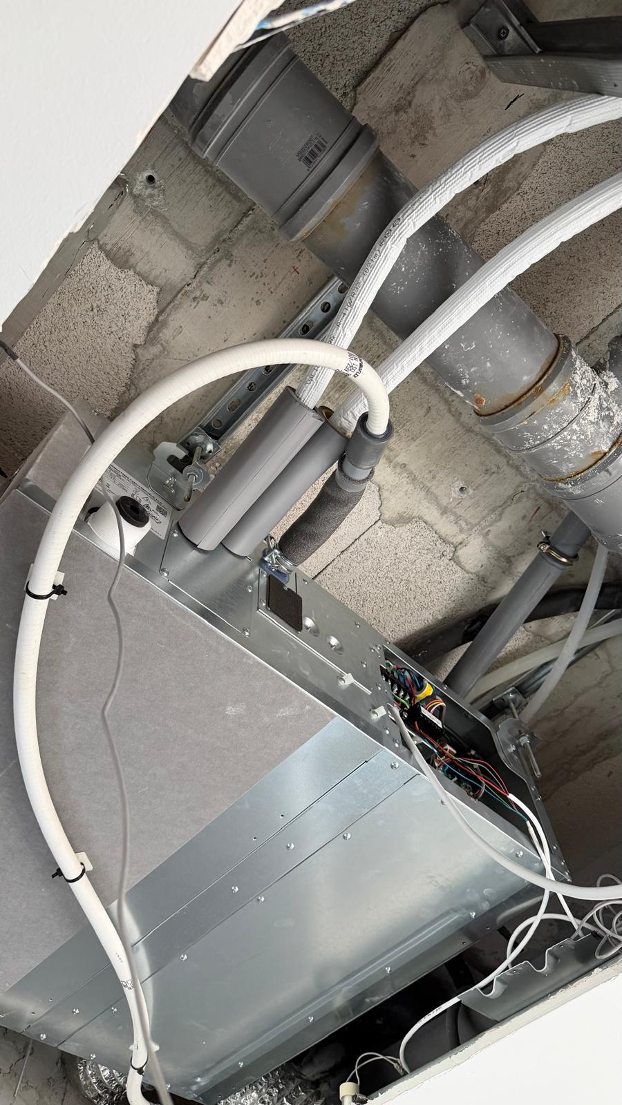 Indoor unit in ceiling void close-up: control board PCB area, condensate drain pipe connections, white refrigerant pipe insulation, concrete block villa ceiling structure