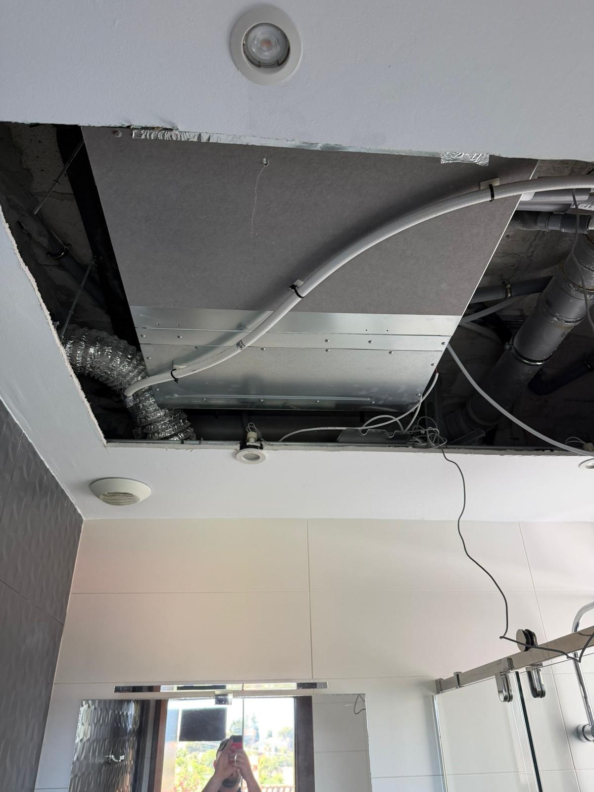 Bathroom ceiling void: unit installed with aluminium flex duct (left), white PVC condensate drain, Airzone control wiring, technician reflected in mirror below