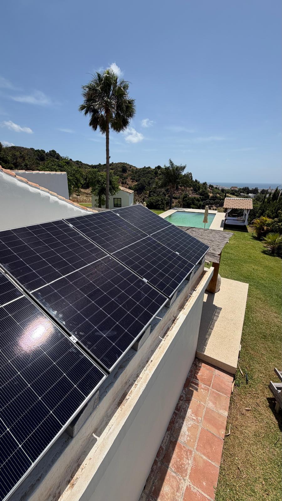 Campo solar en el borde de la cubierta con vistas a la piscina y los jardines, y el mar al fondo — una parcela elevada frente al mar ideal para el autoconsumo.